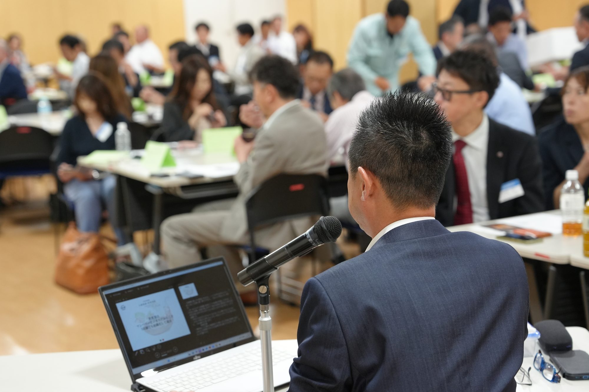 例会でマイクを持ち、参加者に向けて経営報告をする会員の様子